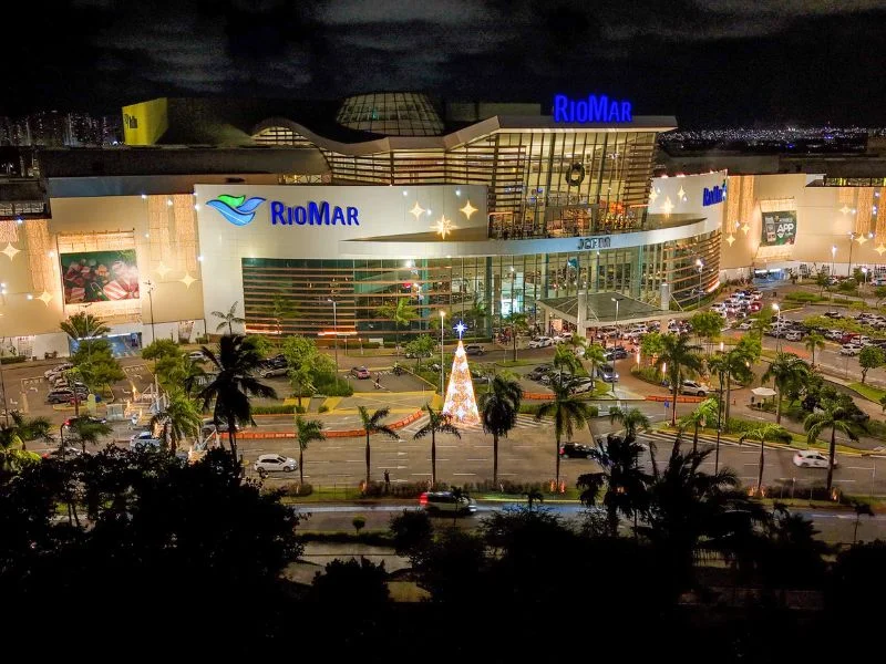 Vista noturna do RioMar Recife com fachada iluminada, carros estacionados, palmeiras e uma árvore de Natal iluminada em frente.