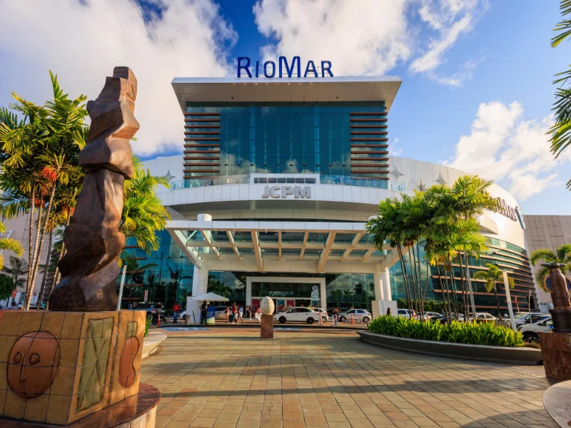 Fachada moderna de um shopping sob céu azul, com grandes painéis de vidro, letreiro "RioMar" no topo e esculturas de arte em destaque no pátio frontal arborizado.