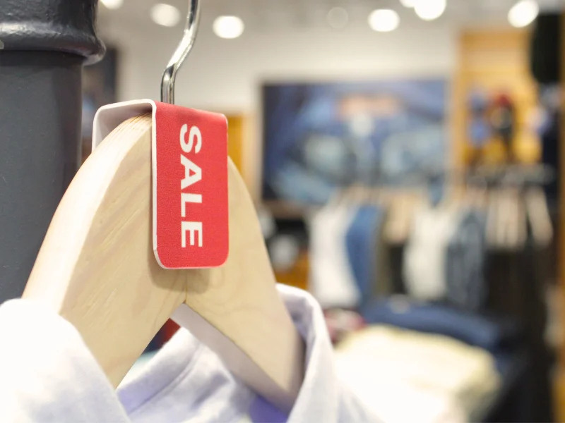 Close em um cabide de madeira sustentando uma peça de roupa clara, com uma etiqueta vermelha de "SALE" em destaque.