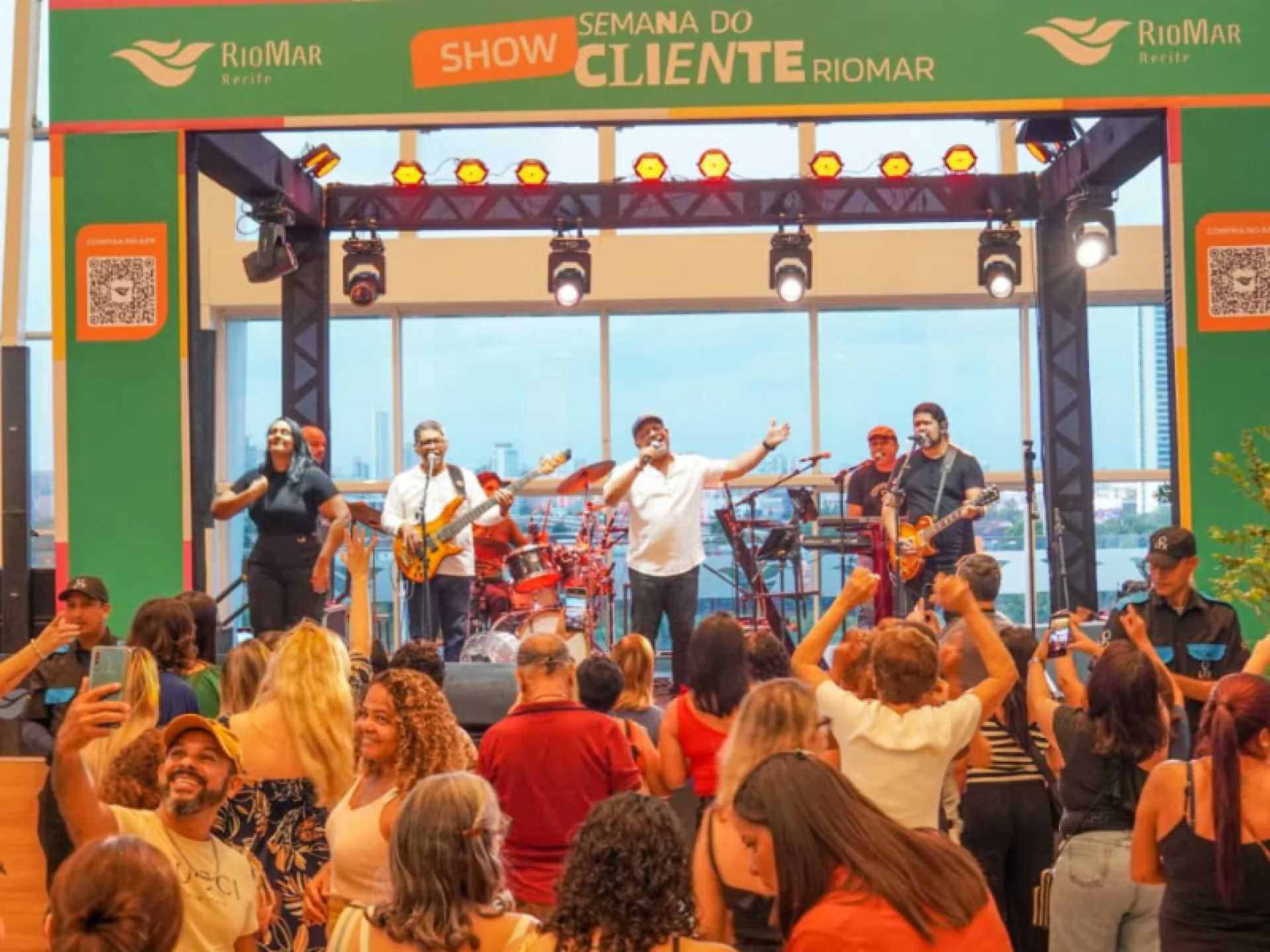 Uma multidão assiste à apresentação de uma banda ao vivo em um palco interno sob uma faixa verde onde se lê "Semana do Cliente". Uma multidão assiste à apresentação de uma banda ao vivo em um palco interno sob uma faixa verde onde se lê "Semana do Cliente".