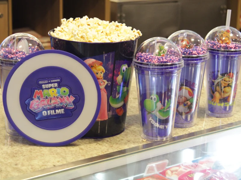 Balde de pipoca cheio e copos roxos temáticos do filme "Super Mario Galaxy" dispostos em um balcão de cinema. Os itens trazem ilustrações coloridas da Princesa Peach, Yoshi, Toad e Bowser.