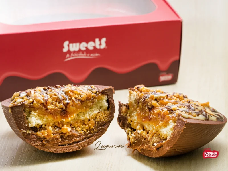 Um ovo de chocolate recheado com camadas de massa clara, caramelo e cobertura crocante, acompanhado por uma caixa vermelha da marca Sweets.