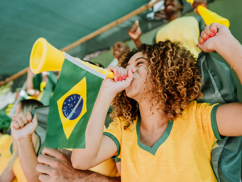 Fotografia em plano médio de uma mulher jovem e sorridente, com cabelos cacheados, vestindo a camisa da seleção brasileira e soprando uma vuvuzela amarela com uma pequena bandeira do Brasil acoplada, cercada por outros torcedores animados.