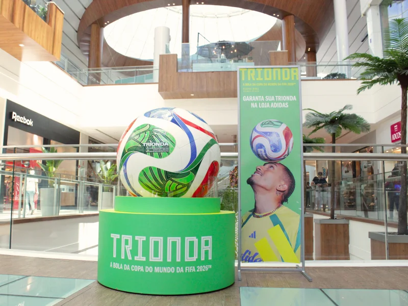 A bola oficial do campeonato mundial de futebol de 2026, a Trionda, está em exposição no centro do hall de um shopping (mall). A bola está posicionada sobre uma grande base cilíndrica verde-limão, que leva o nome "TRIONDA".