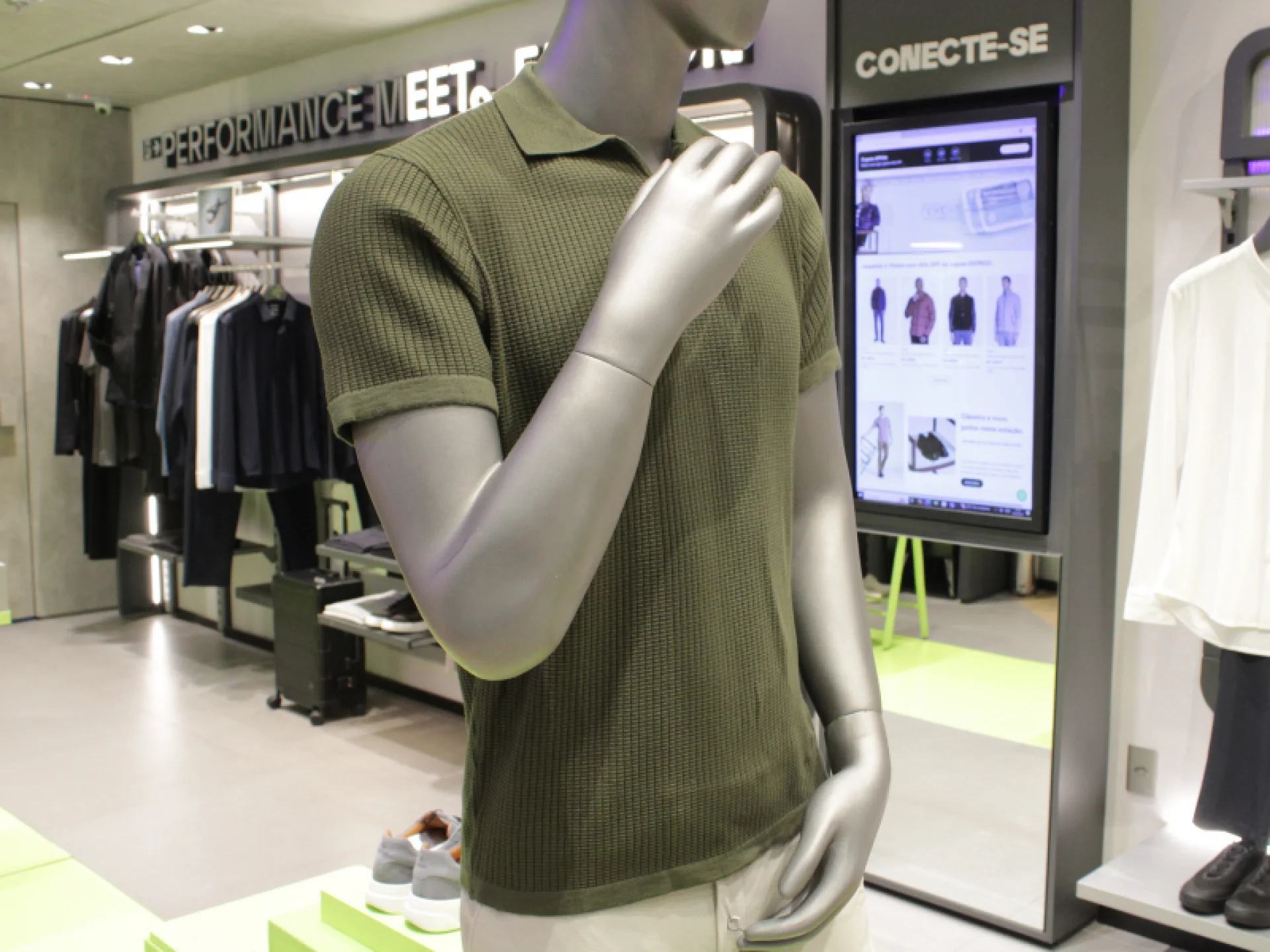 A imagem foca no torso de um manequim vestindo uma polo de malha canelada ou texturizada na cor verde-militar, combinada com uma calça clara.