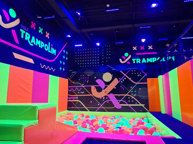 Área interna do parque sob luz negra, destacando uma piscina de blocos de espuma coloridos, paredes acolchoadas em neon e o logotipo do parque ao fundo.