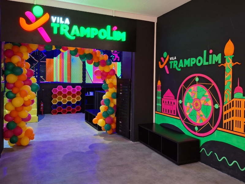 Entrada de um parque de trampolins com arco de balões coloridos, letreiro luminoso neon "Vila Trampolim" e paredes pretas decoradas com grafites vibrantes.