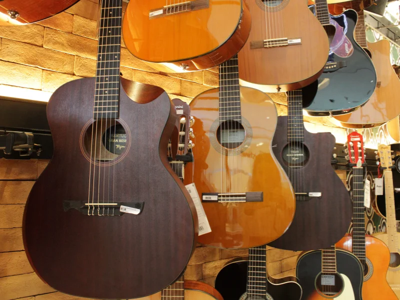 Vários violões acústicos de diferentes tons de madeira e cores (claro, escuro e preto) pendurados lado a lado em uma parede de tijolos claros. O destaque frontal é um violão de madeira fosca escura com corte cutaway.