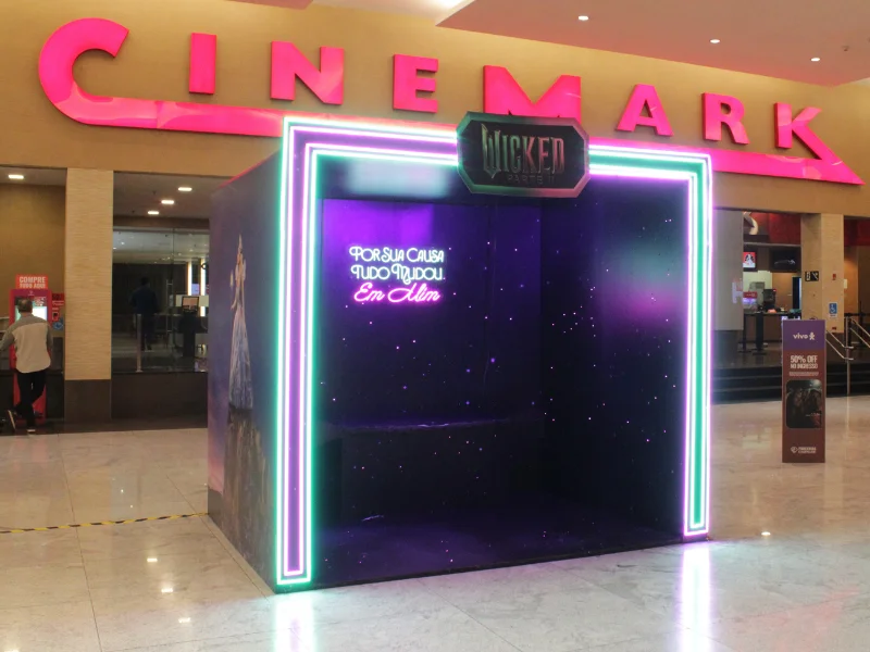 Visão frontal da cabine promocional "Wicked Parte II" no cinema, com o letreiro Cinemark em rosa ao fundo. A cabine tem iluminação neon e um interior escuro com estrelas brilhantes.